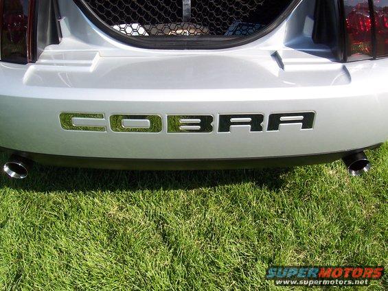 Rear bumper of a silver Ford Mustang with 'COBRA' lettering and dual exhaust pipes