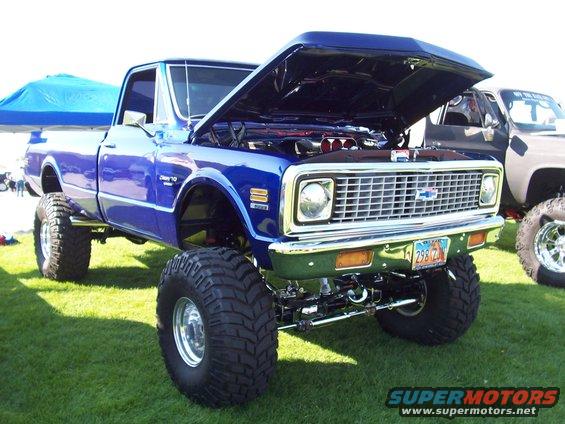 Blue lifted pickup truck with hood open, showing engine, large tires, and chrome grille