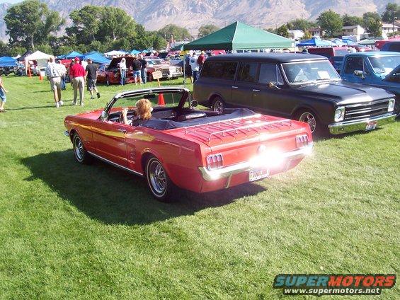 Red convertible with a woman driving, parked on grass at an outdoor event with other cars and tents