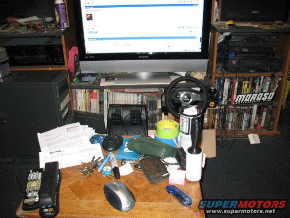 Desk with computer, gaming wheel, pedals, remotes, keys, and papers