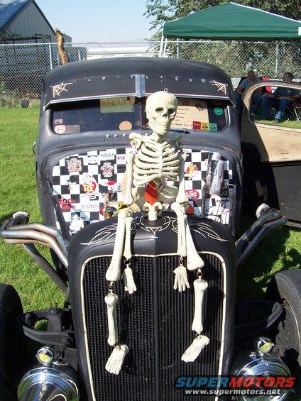 Black hot rod with a skeleton sitting on the hood, checkered flag and stickers on the grille area, and chrome exhaust pipes
