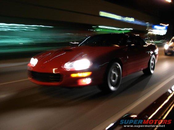 Red sports car with headlights on, driving at night. Visible text: SUPERMOTORS.NET
