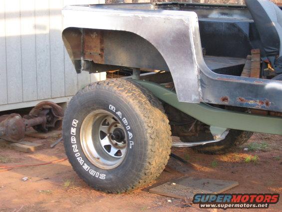 Jeep CJ body with rear fender removed, showing rust and unfinished metal. A spare axle sits nearby