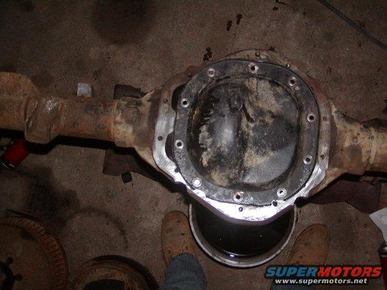 Differential axle housing with cover removed, sitting over a bucket of dark fluid. Rusty metal and concrete floor visible