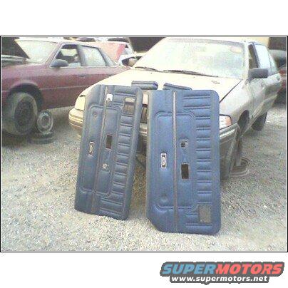 Two blue car door panels with textured inserts and chrome trim, resting on gravel in front of a junkyard car