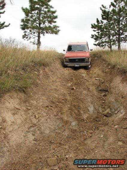 Orange van going up a steep, rutted dirt hill with trees and dry grass around