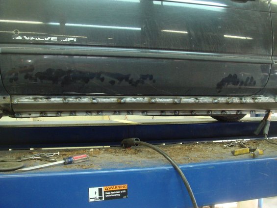 Dark colored vehicle's rocker panel with rust and welding repairs, viewed from the side