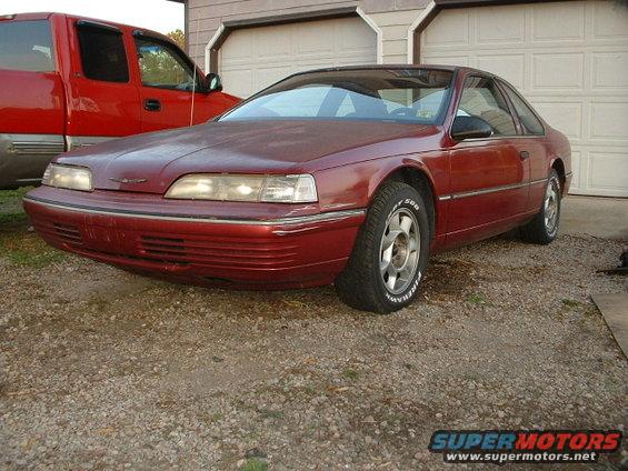Red 1989 Ford Thunderbird coupe parked outside, with a red pickup truck to its left