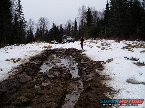 Off-road vehicle navigating a challenging muddy and snowy terrain with a person standing nearby