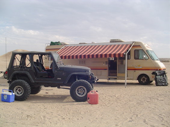 Black Jeep TJ with aftermarket wheels and tires, parked next to a tan RV with red striped awning. Red gas can and blue cooler nearby