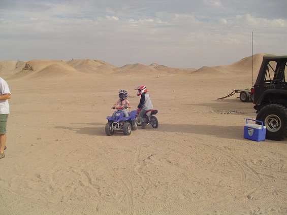 Two kids in helmets riding ATVs and dirt bikes in a sandy desert landscape with a Jeep and trailer in the background