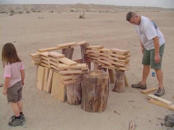 Child and man in desert building a structure with wood planks and logs
