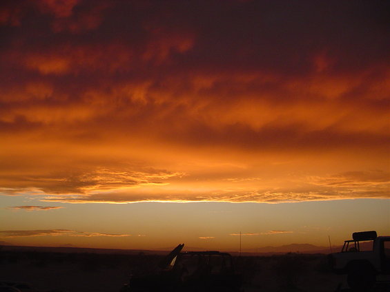 Sunset sky with orange and red clouds over a desert landscape. Silhouettes of vehicles are visible in the foreground