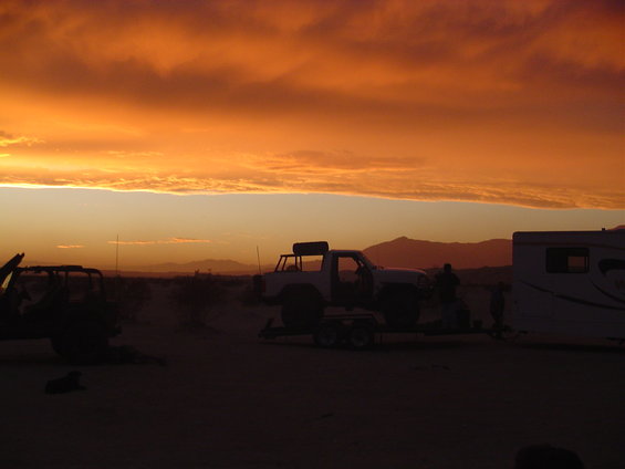 White Jeep Wrangler on a trailer at sunset, with a camper and another off-road vehicle visible