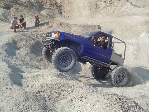 Blue pickup truck with off-road modifications climbing a dirt hill. Driver is visible. Three people watch from the side