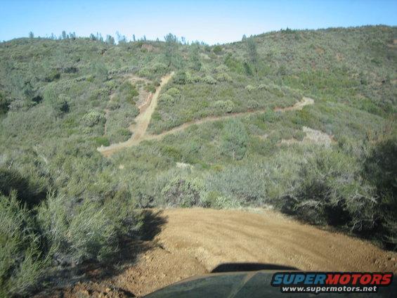 Dirt trail winding up a scrub-covered hill under a clear blue sky. Part of a vehicle's hood is visible in the foreground