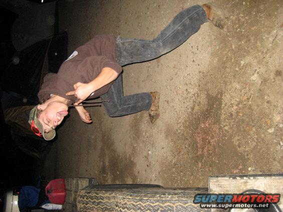 Person in jeans and hoodie making a rude gesture, lying on the ground next to a tire and gas can