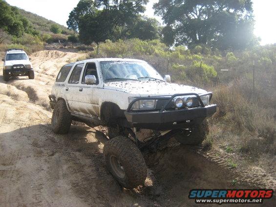 White Toyota 4Runner with aftermarket bumper and large tires navigating a dirt trail, another white SUV behind it
