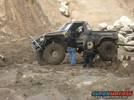 Muddy off-road truck with roll cage and large tires, two people standing nearby