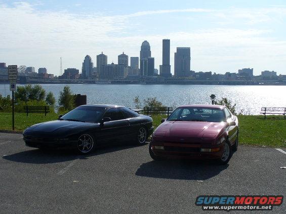 Two 1991 Ford Probes parked by a river with a city skyline