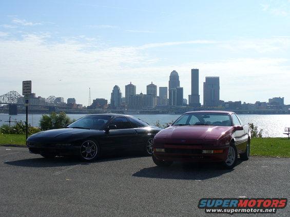 Two 1991 Ford Probes parked near a river with a city skyline. One is black, the other is red