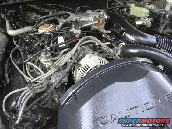 Engine bay with visible wiring, alternator, and black air intake. CAUTION text on cover