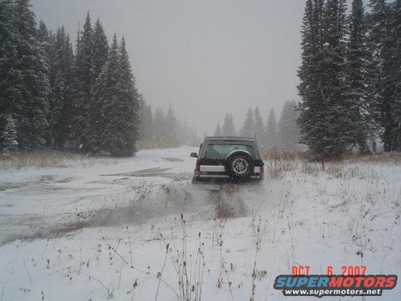 Dark SUV with spare tire on rear door driving on a snowy road surrounded by pine trees