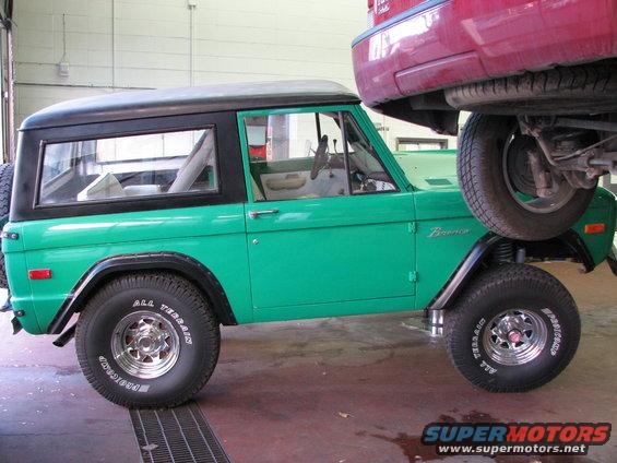 Green 1975 Ford Bronco with a black top, viewed from the side, with a red car lifted above it