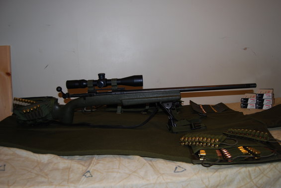 Green rifle with scope and bipod, next to ammo pouches and boxes