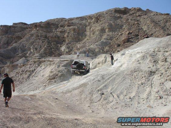 White SUV with spare tire on rear door, driving on a dirt hill with people around