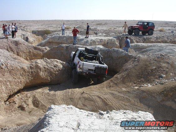 White off-road SUV with spare tires on roof rack navigating a dirt canyon, red Jeep in background