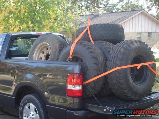 Dark gray pickup truck bed loaded with four large off-road tires secured by orange straps