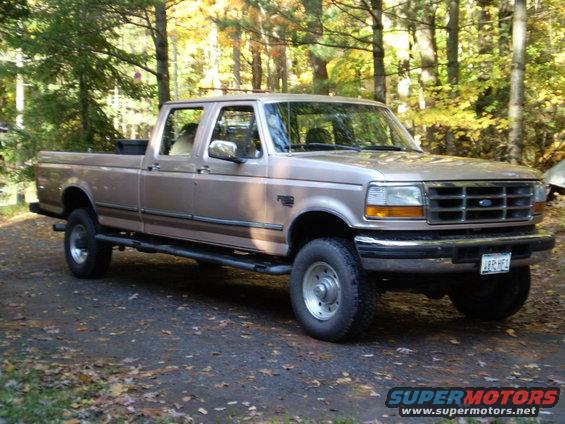 Tan Ford F-Series truck with a crew cab and long bed, parked on a dirt road with fallen leaves. The grille and headlights are visible