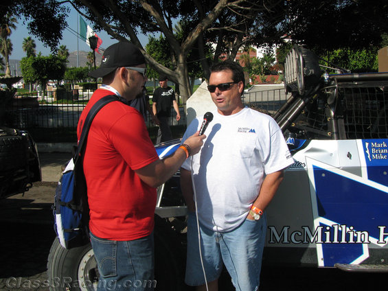 Man in white shirt with 'McMillin Racing' logo is interviewed next to a custom off-road race truck