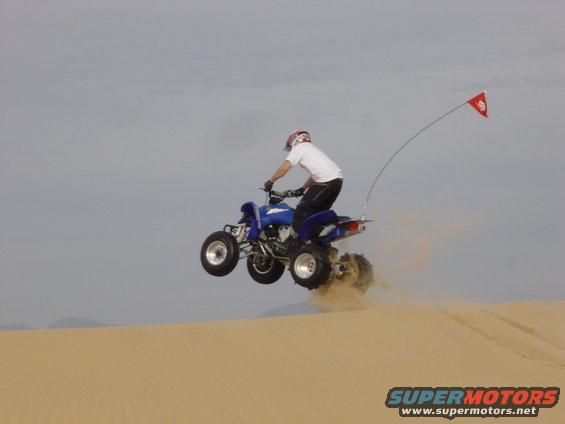 Blue ATV airborne over sand dune with red flag