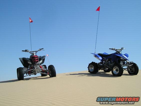 Two quad bikes with red flags on sand dunes under a clear blue sky