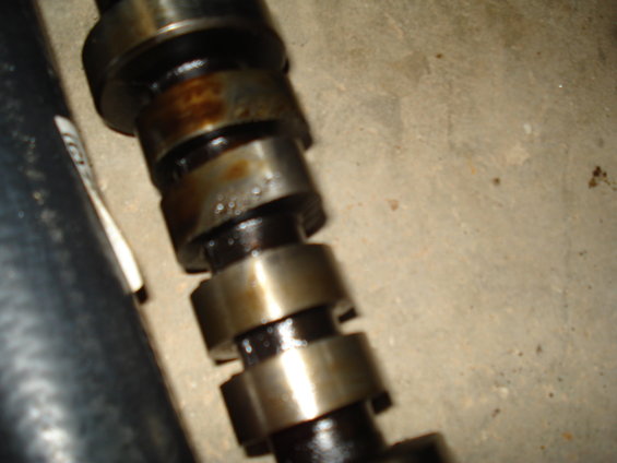 Camshaft with oil residue and metal lobes on a concrete surface next to a black hose