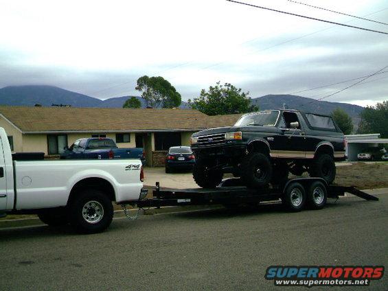 Black lifted Ford Bronco on a trailer being towed by a white Ford F-250. Another blue truck and a dark sedan are parked in the driveway