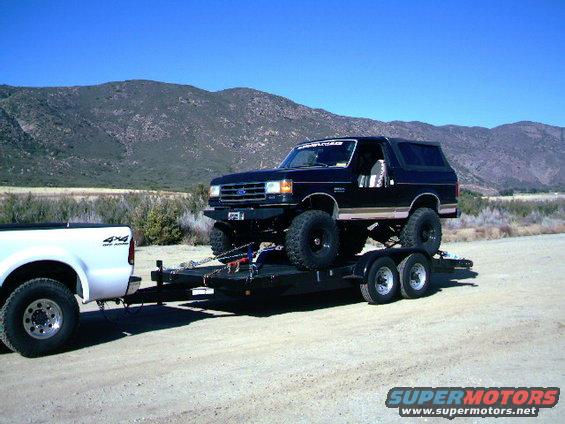 Black lifted Ford Bronco on a trailer, towed by a white Ford F-250 pickup truck. Mountains in background