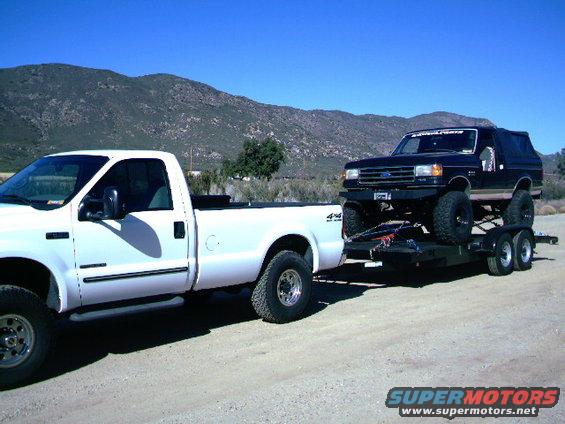 White Ford pickup truck towing a black Ford Bronco on a trailer, side view