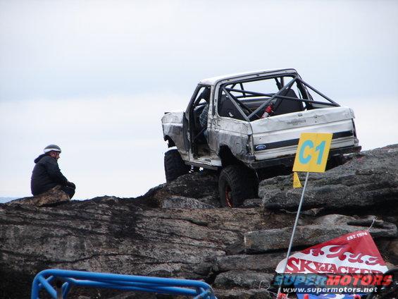 Modified SUV with roll cage on rocky terrain, person sitting on rock nearby