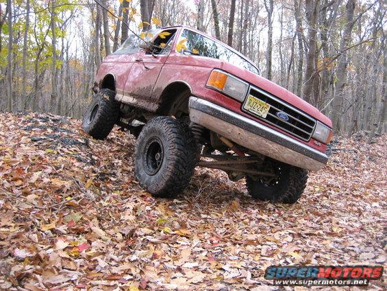 1990 Ford Bronco with lifted suspension and large tires driving off-road on a leaf-covered hill