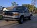 1995 Ford F-150 - This two-tone Ford F-150 pickup truck is parked on a paved surface with a residential building and tropical foliage behind it.