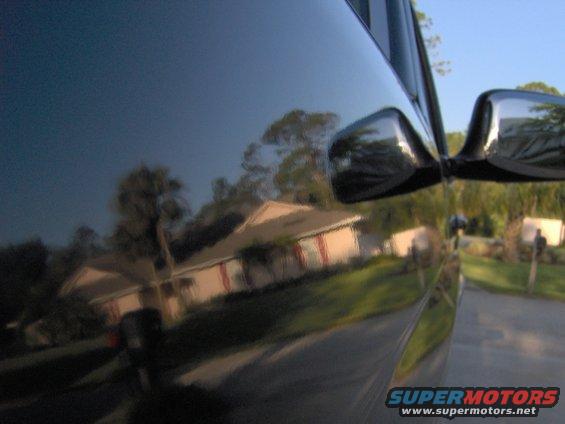 Black truck side mirror reflecting a house and trees. Watermark "SUPERMOTORS" visible