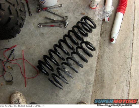 Two black coil springs and a red-topped shock absorber lie on a concrete floor next to a tire and tools