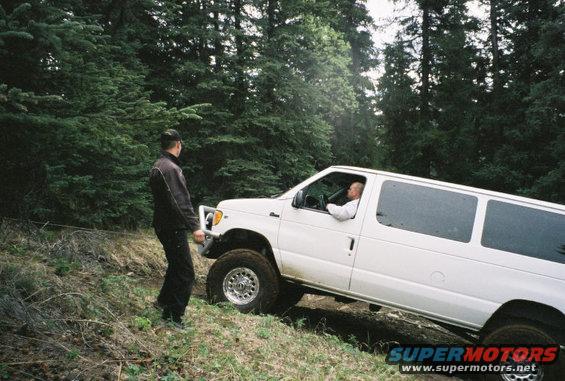 White Ford E350 van with off-road tires on a dirt path, driver visible inside