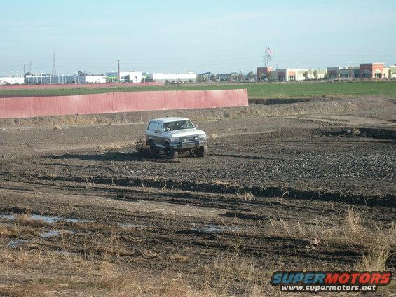 1989 Ford Bronco driving through a muddy field with buildings in the background