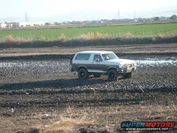 Two-tone Ford Bronco driving through a muddy, rocky area with fields and buildings in the background