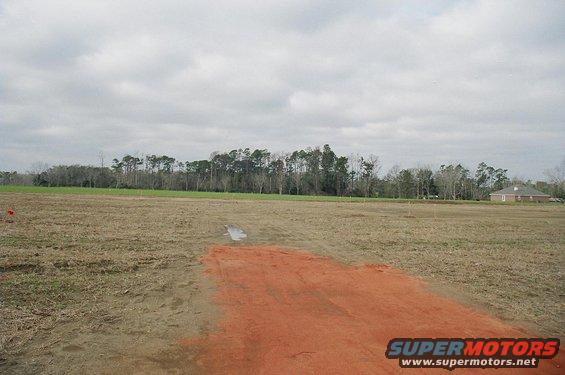 Construction site with bare ground, orange dirt, and trees in the background under a cloudy sky. A house is visible on the right