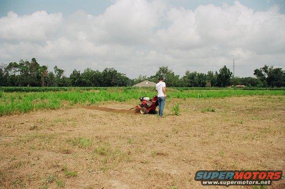 Person operating a tiller in a field with trees and a building in the background